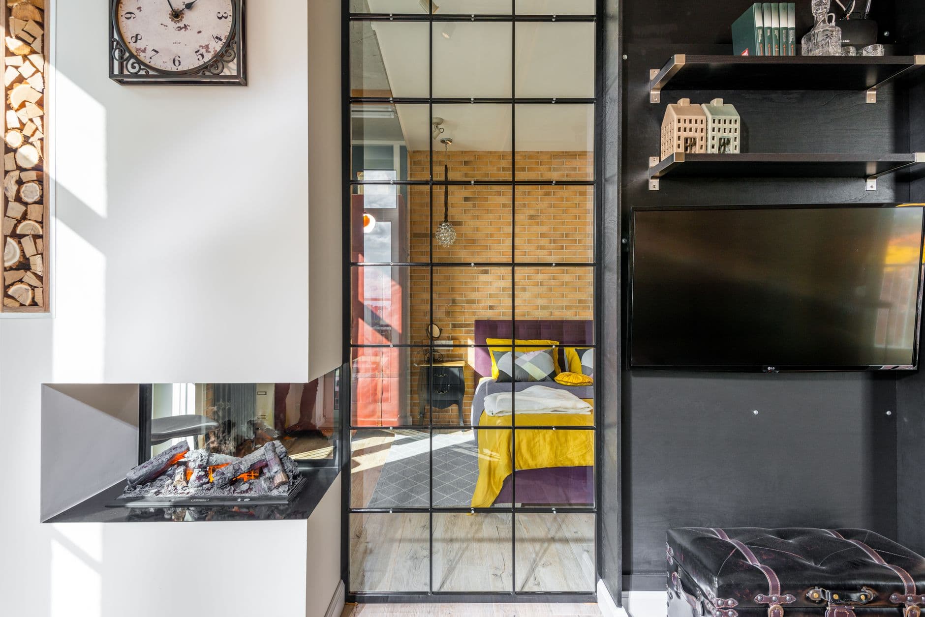 Colorful bed with cushions behind glass doorway seen from light modern room with fireplace and shelves with decorations at home